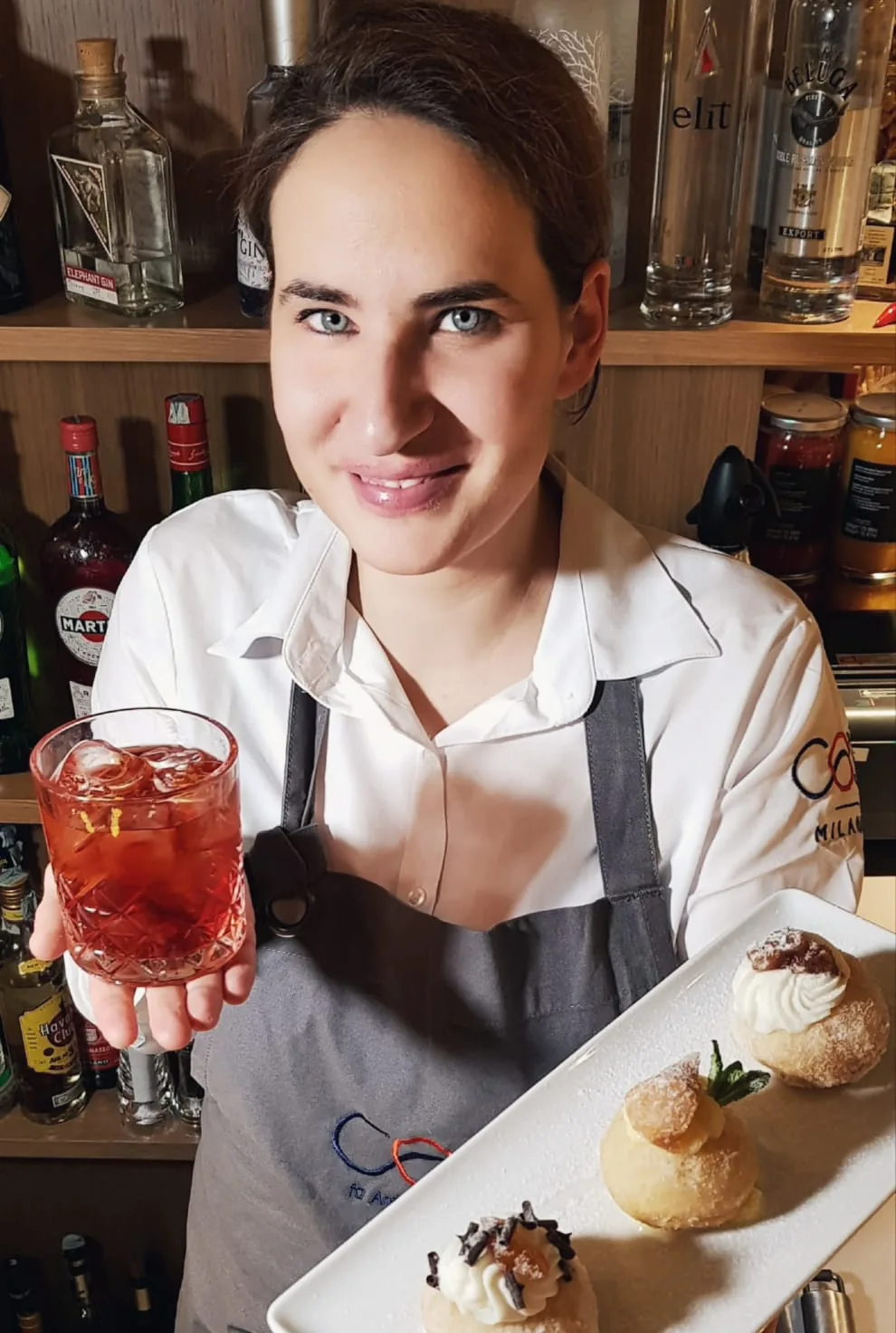 Rita dietro il bancone con un cocktail e un vassoio di dolci